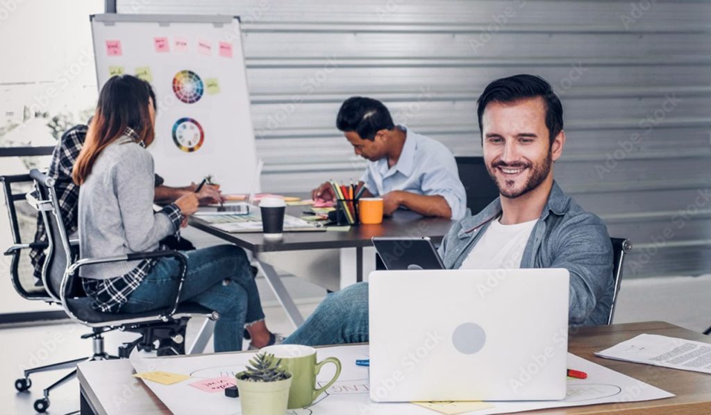 In the modern office, the director of creative design is reviewing work on his laptop as the crew is casually meeting in the background.